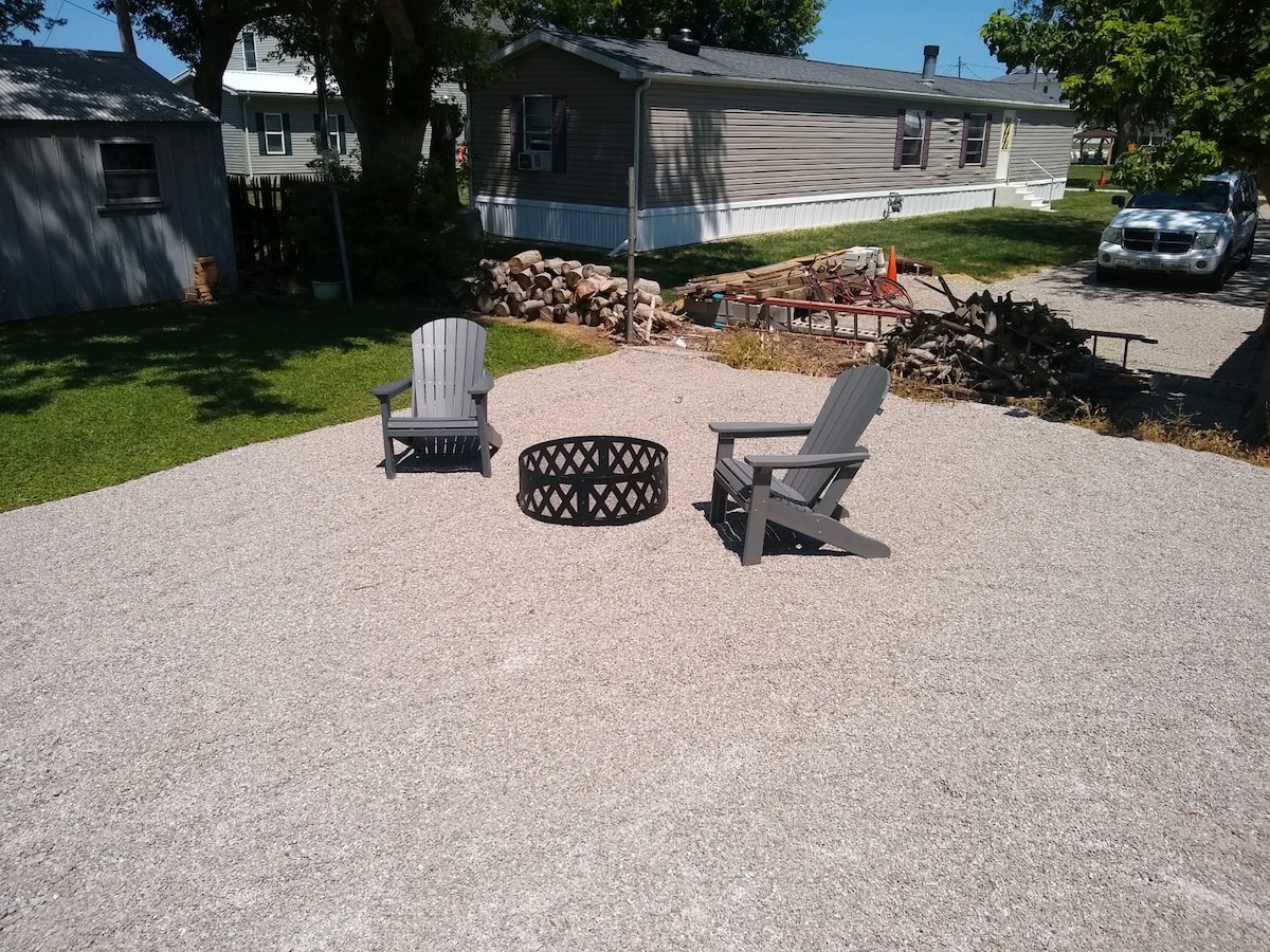 A gravel seating area features two Adirondack chairs positioned around a metal fire ring. The surrounding space is clear and offers easy access to nearby greenery and structures, creating a relaxed outdoor spot for gatherings.