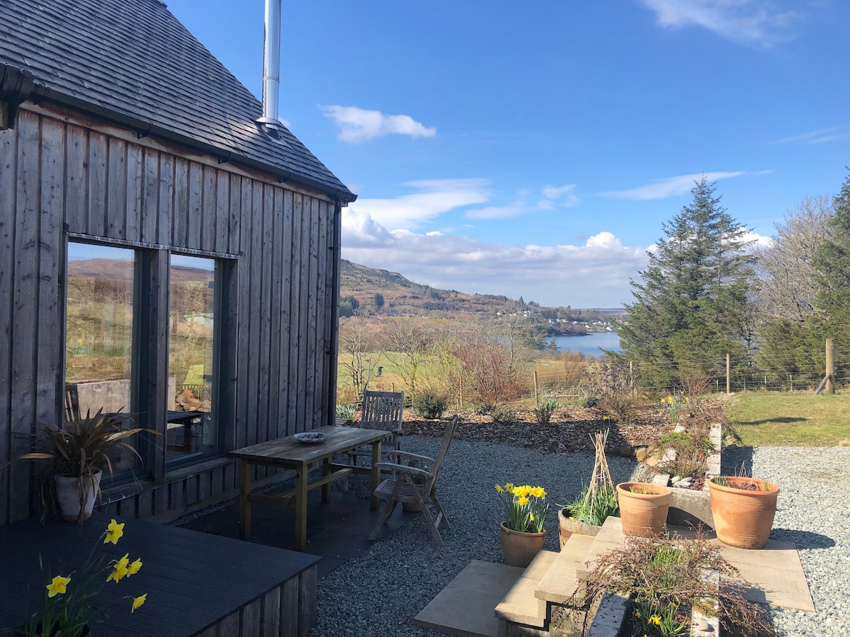 A contemporary larch wood house is depicted, with a sunny patio area featuring potted flowers and a table with chairs. Scenic views of the lush landscape and water are visible in the background, enhancing the tranquil outdoor space.