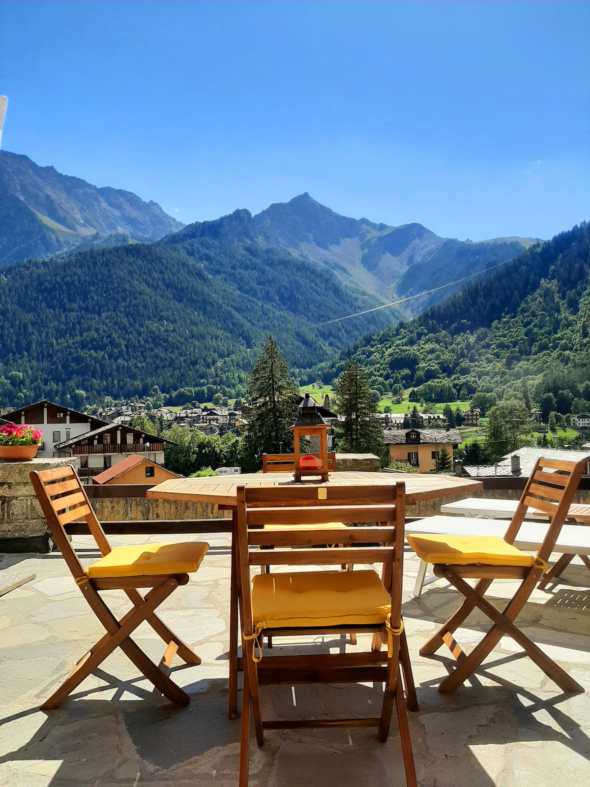 A terrace area is shown with four wooden chairs, featuring yellow cushions. In the background, expansive mountain views surround the scene, highlighting lush greenery and a clear blue sky. The setting suggests a peaceful outdoor space for relaxation or dining.
