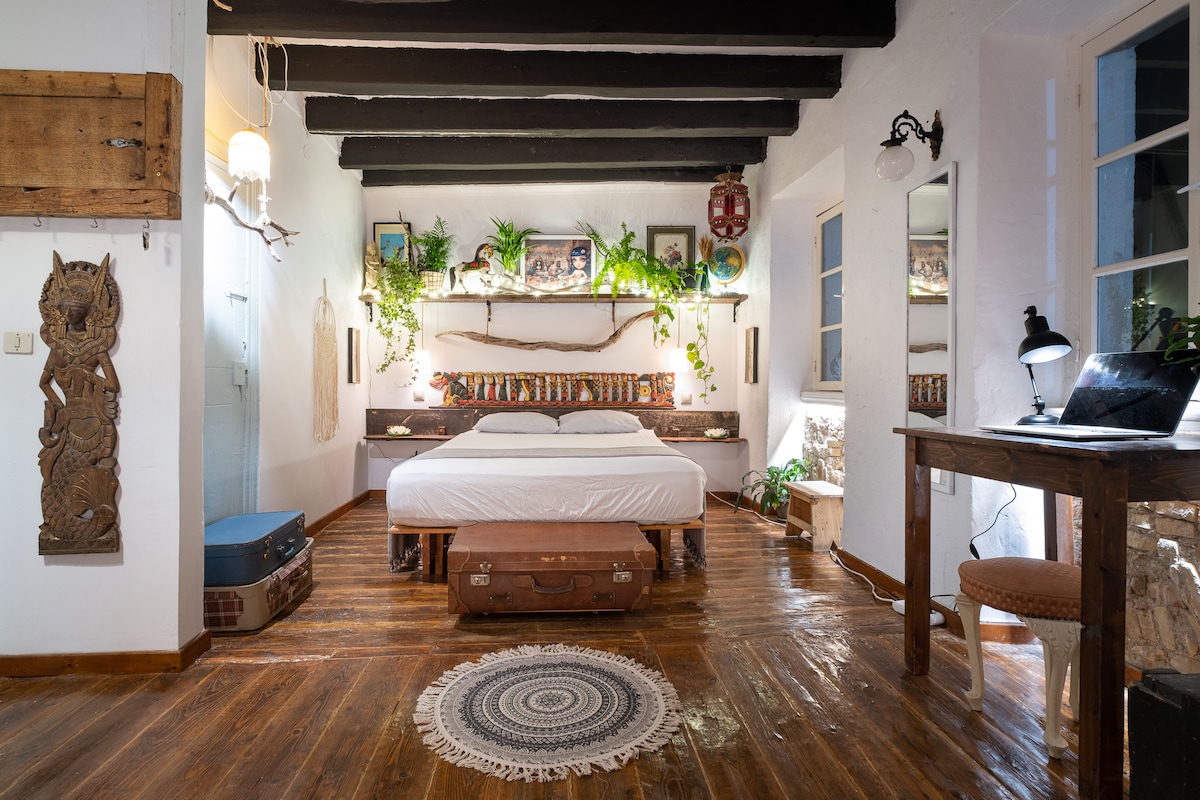 A spacious bedroom features dark wooden beams and warm wooden flooring. A large bed is positioned against a decorative wall adorned with plants and art, while a desk and lamp are placed nearby. Soft lighting creates a serene environment, contributing to a cozy atmosphere.