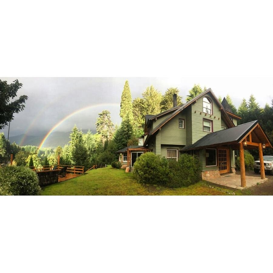A three-story house surrounded by lush greenery features a prominent porch with a wooden overhang. A vibrant rainbow arcs across the sky, enhancing the natural scenery. Trees line the property, offering a serene setting in this tranquil landscape.