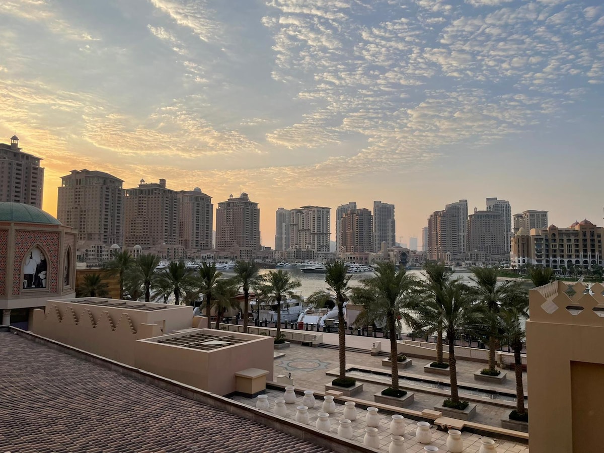 A wide view of a Marina at sunrise shows buildings along the waterfront, framed by palm trees. The sky features soft clouds in warm tones, creating a serene atmosphere. The foreground includes a well-maintained plaza with smooth pathways, enhancing the inviting setting.