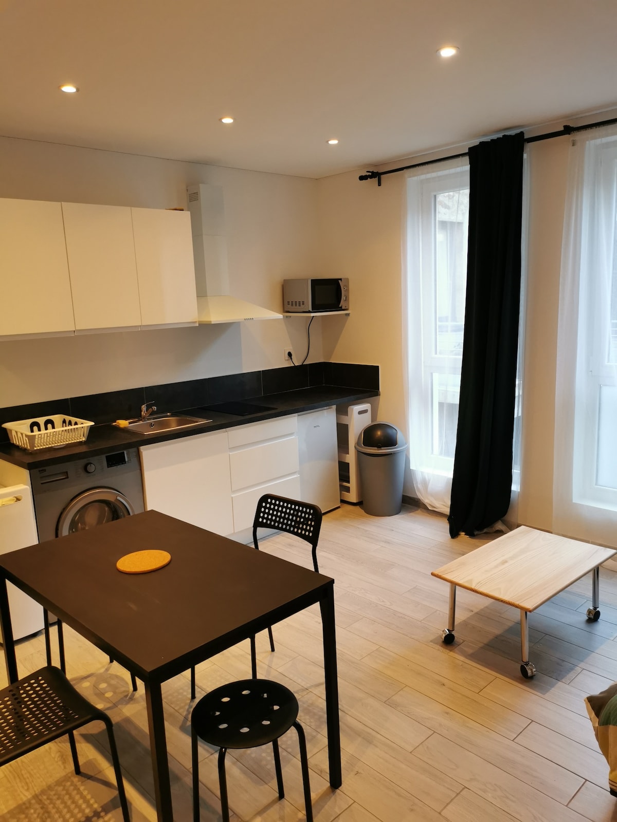 The kitchen area features sleek white cabinets with a black countertop. A round wooden table is surrounded by four black chairs. A washing machine sits underneath the counter, and a window allows natural light to brighten the space. A small wooden cart is positioned in front.