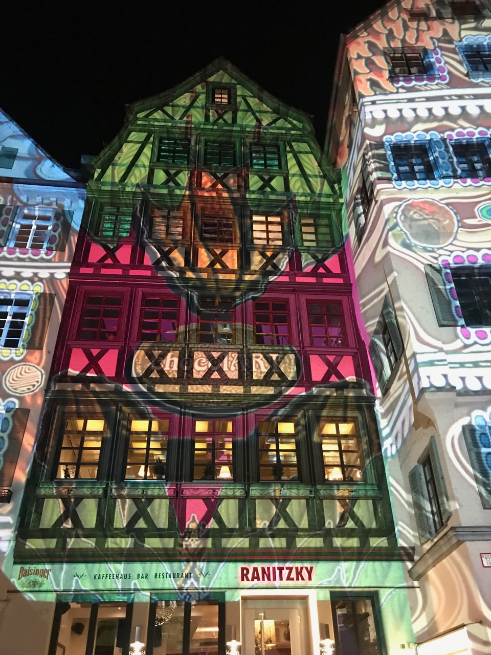 An historic building’s façade features vibrant, colorful projections that illuminate its architectural details. The graphic designs playfully enhance the traditional elements, creating a dynamic visual display. Large windows and distinct patterns are prominent against the dark night backdrop.