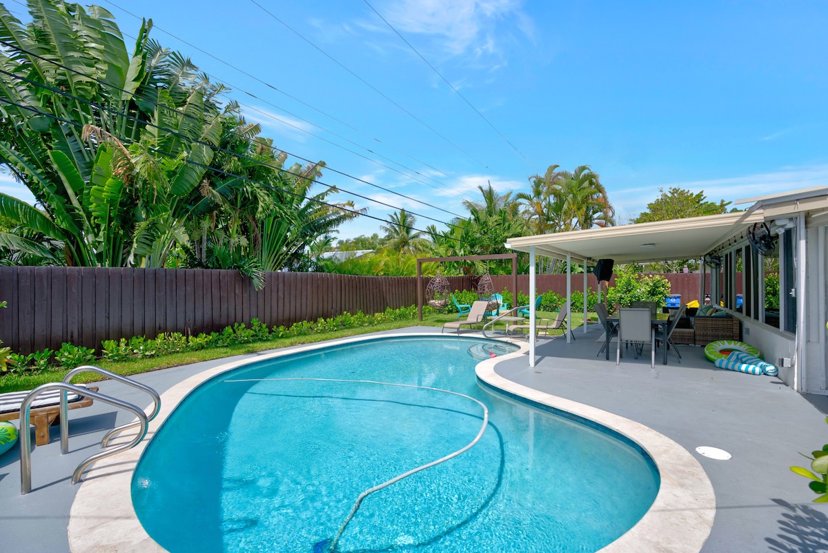 A spacious, inviting swimming pool is surrounded by a landscaped backyard, featuring lush greenery and palm trees. Lounge chairs and a shaded seating area are positioned for relaxation. The poolside area is designed for enjoyment, perfect for soaking up the sun or unwinding in the shade.