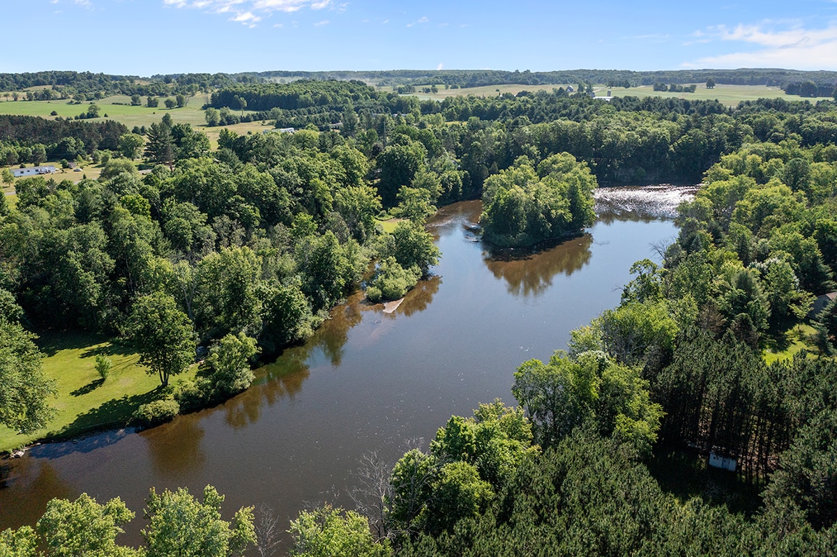 A scenic view captures the winding river surrounded by lush greenery. Trees line the riverbanks, offering a vibrant mix of foliage. Open fields extend beyond, contributing to a serene and natural landscape under a clear blue sky.