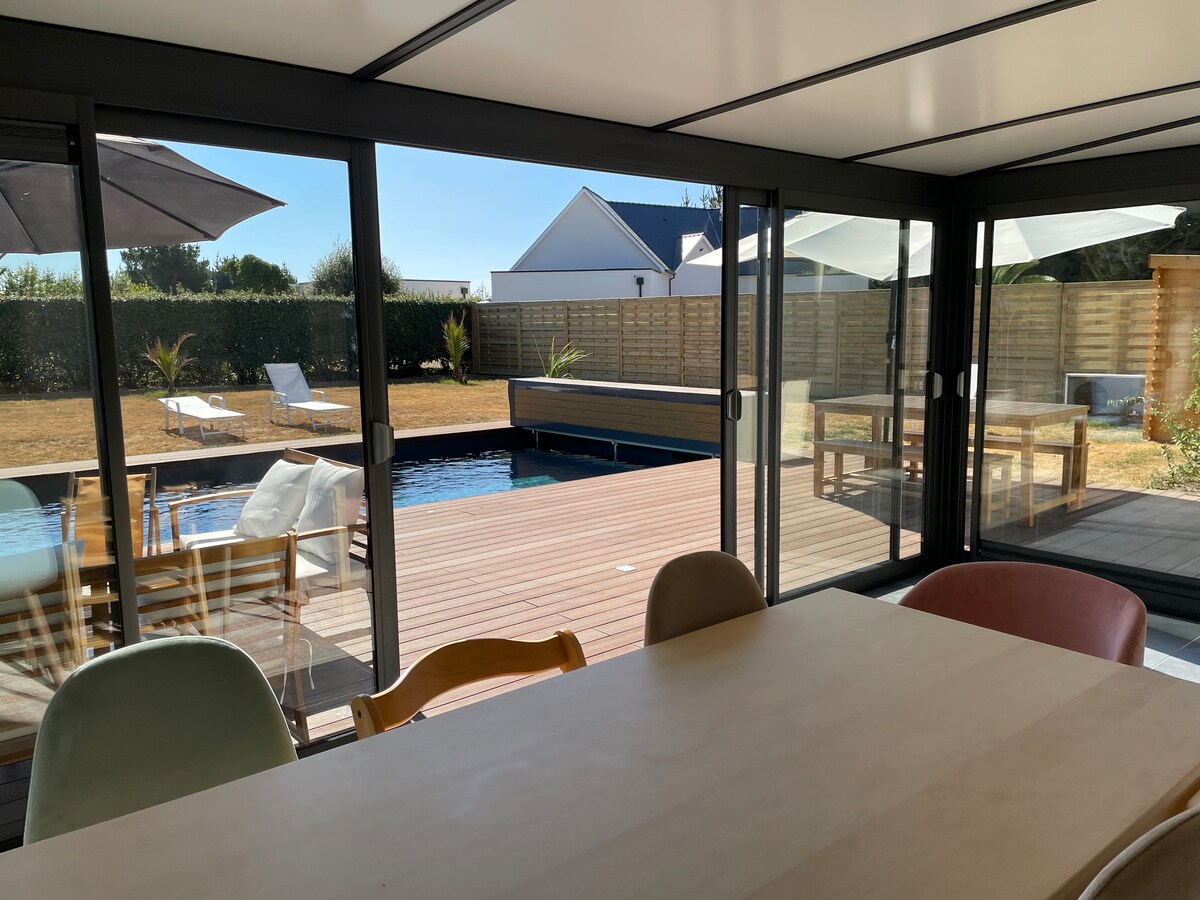 A well-lit space features large glass doors that open to a wooden terrace. Outdoor seating and lounge chairs overlook a heated pool. The dining area includes a large table surrounded by colorful chairs, creating an inviting atmosphere for gatherings.