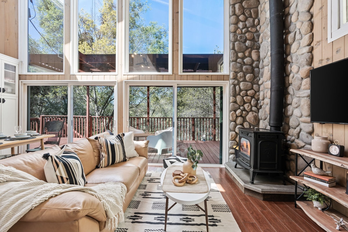 A spacious living area features large windows that allow natural light to flood in, with views of the surrounding trees. A stone fireplace is positioned beside a comfortable, neutral-toned sofa adorned with decorative cushions. A round coffee table sits in the center, adding warmth to the inviting space.