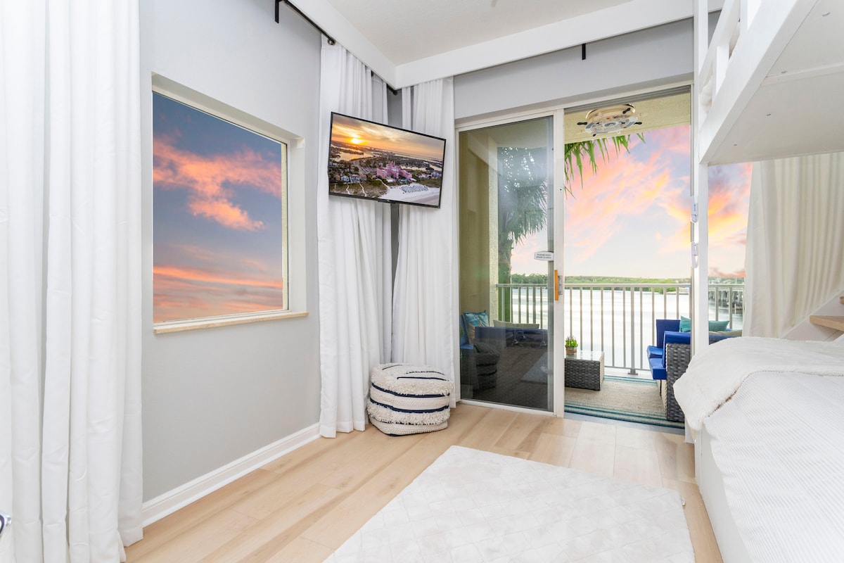 A light-filled room features large windows offering views of a colorful sunset. A flat-screen TV is mounted on the wall opposite a cozy sleeping area with a bunk bed. Soft curtains frame the windows, enhancing the serene atmosphere.