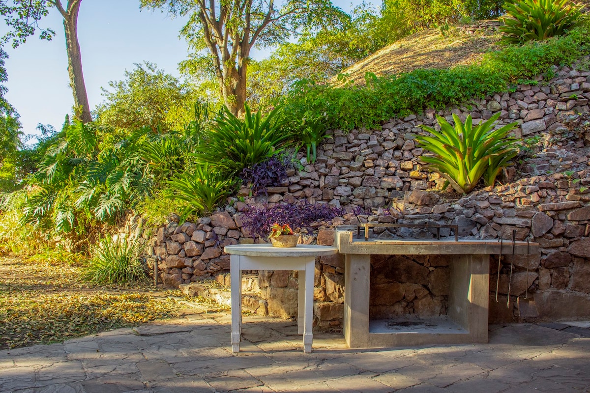 An outdoor grilling area is featured, with a stone backdrop and lush greenery surrounding the space. A rustic table stands alongside a concrete grill, providing a functional area for cooking. The ground is paved with stones, and dappled sunlight filters through the trees.
