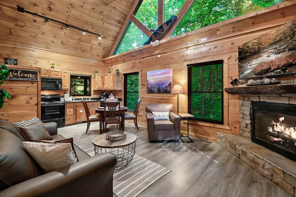 A cozy living area features a stone fireplace, complemented by a comfortable sofa and armchair. Natural light filters through green-tinted blinds, enhancing the wooden interior. A dining table for four is positioned nearby, creating a welcoming space for relaxation and meals.