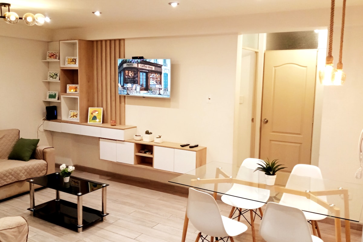 A modern living area features a light-colored couch, a sleek glass coffee table, and a dining table surrounded by white chairs. A mounted television displays an urban scene. Decorative shelves showcase colorful accents, and soft lighting enhances the welcoming atmosphere.