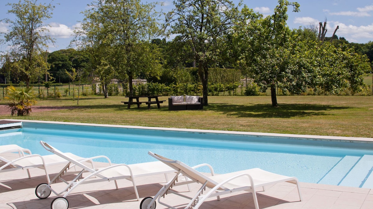 The image features a clear swimming pool surrounded by several white lounge chairs. Lush green trees and a grassy area are visible in the background, along with a seating area consisting of a couch and picnic table, providing a serene outdoor setting.