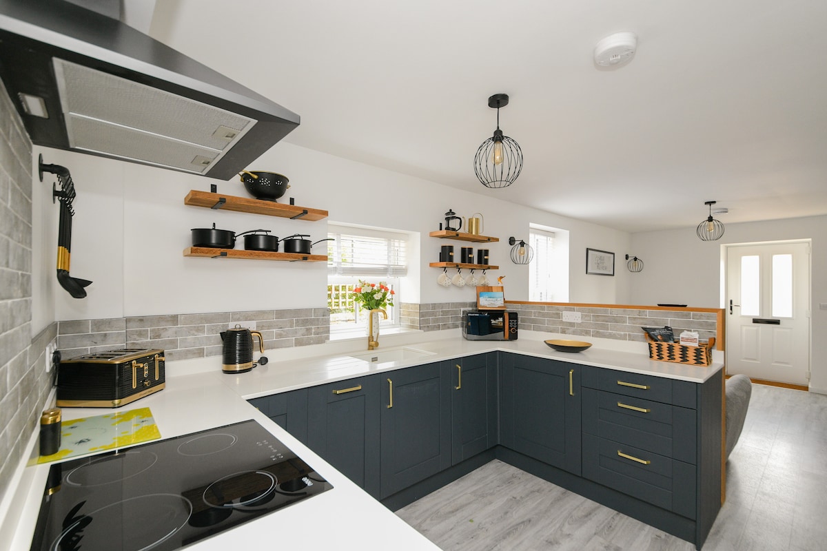 The kitchen is designed with modern fixtures and a functional layout. Dark cabinetry and light countertops provide a contrast. Open shelving displays cooking utensils and decorative items. Natural light enters through a window, illuminating the space. An entrance door is visible in the background.