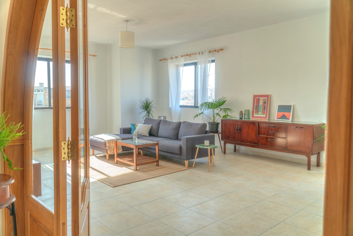 A spacious living room is visible, featuring a large gray sectional sofa and a wooden coffee table. Natural light enters through large windows, illuminating a neutral-toned area rug. A wooden sideboard displays artwork and plants, creating a relaxed ambiance in the corner.