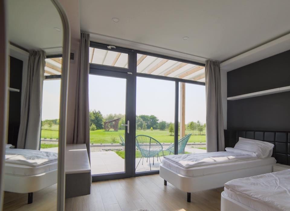 A bright room features two spacious beds positioned beside large glass doors, leading to an outdoor seating area. Natural light fills the space, while soft curtains gently frame the windows. A mirror reflects the serene view of the green landscape outside.