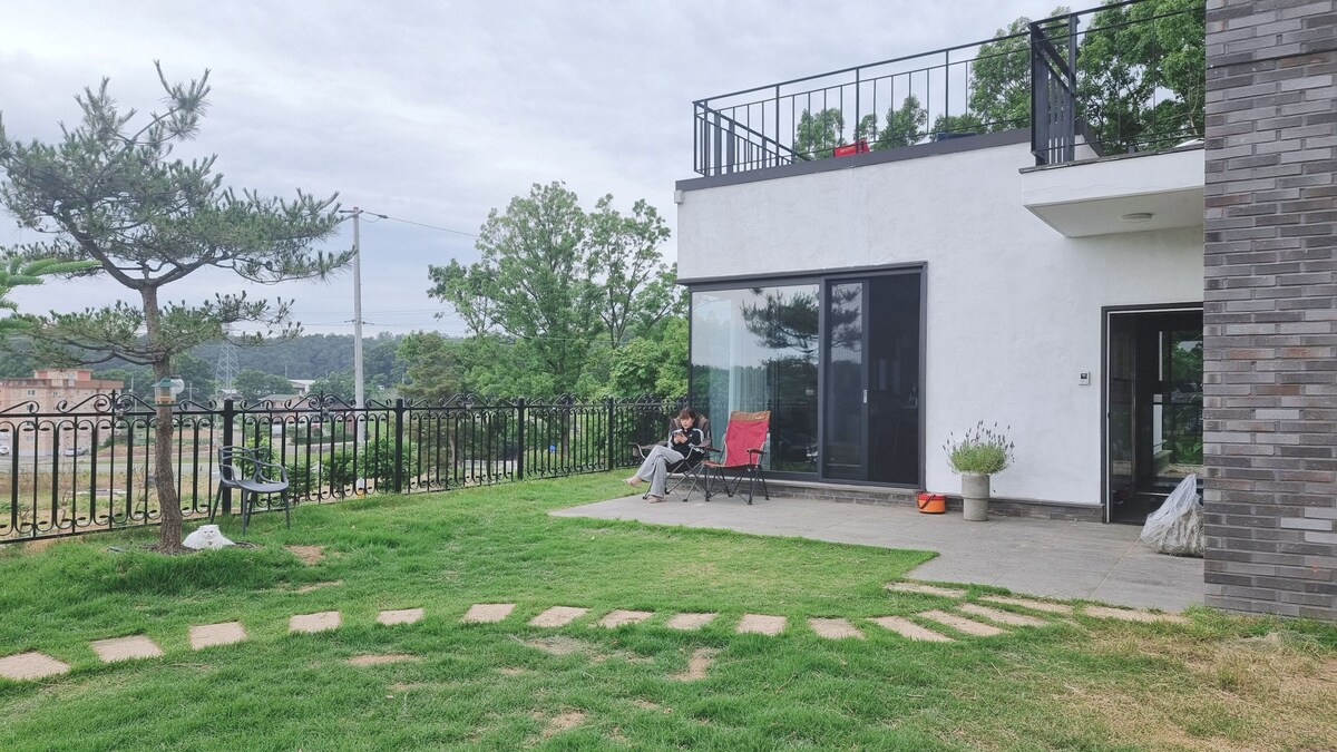 A spacious outdoor area features a well-maintained lawn bordered by low stone pathways. The modern two-story house includes large glass windows, showcasing views of the surrounding greenery. A patio area is present, with two chairs comfortably arranged for relaxation.