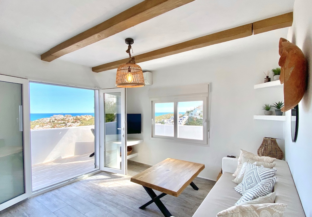 A welcoming lounge area features large glass doors opening to a terrace with sea views. A simple wooden coffee table is centered in front of a sectional sofa adorned with decorative pillows. A woven light fixture hangs above, while wall shelves display small plants.