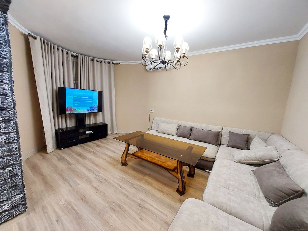 A spacious living area features a large, light-colored sectional sofa arranged around a wooden coffee table. A flat-screen television is mounted on a wall opposite the seating. Soft, neutral curtains frame the windows, enhancing the bright and open feel of the room.