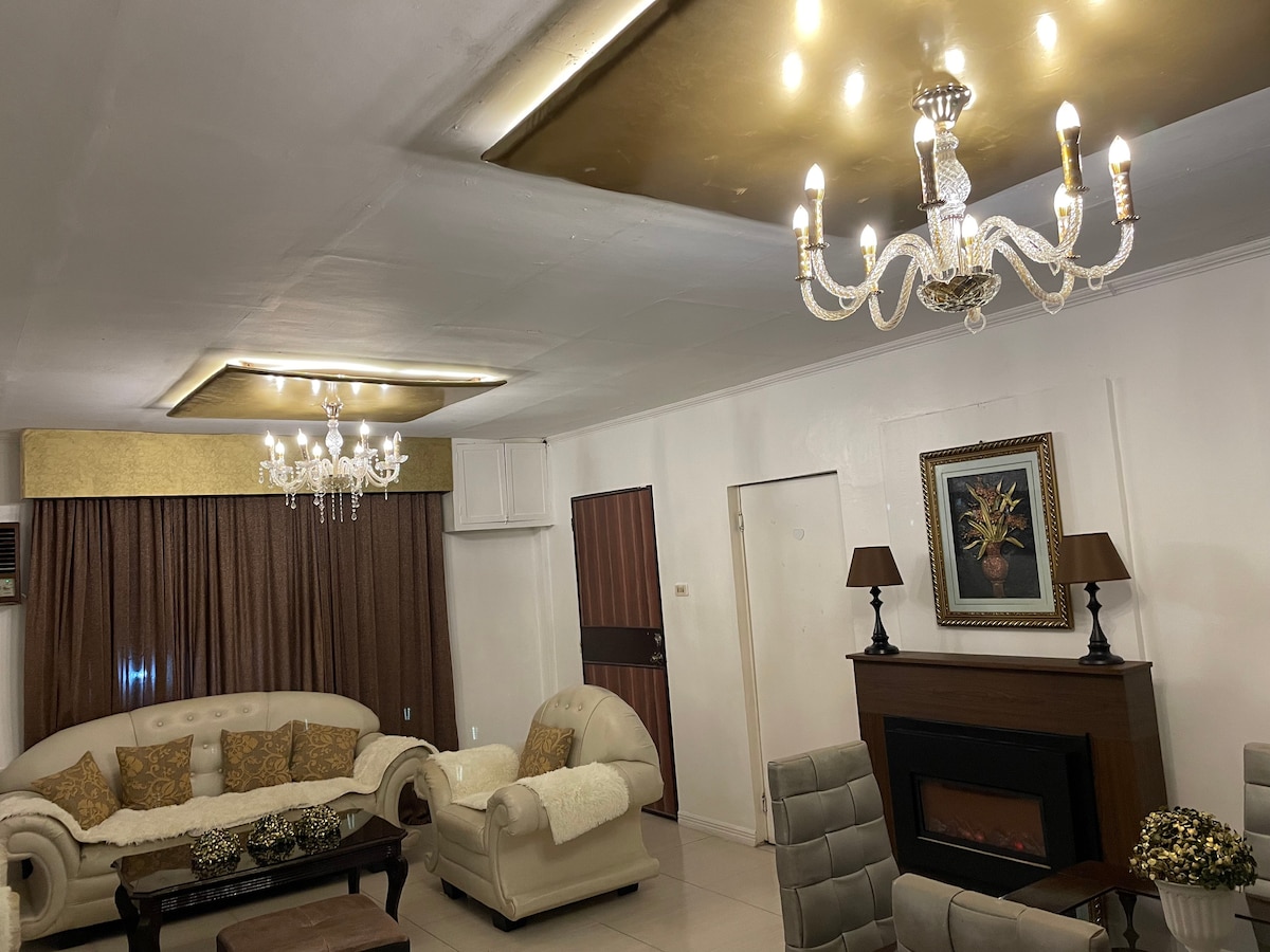 A spacious living area is featured, showcasing a light-colored sofa with decorative cushions, complemented by a stylish coffee table. Two elegant chandeliers provide bright lighting, while warm tones are carried through the curtains and wall decor, creating a comfortable and inviting space.
