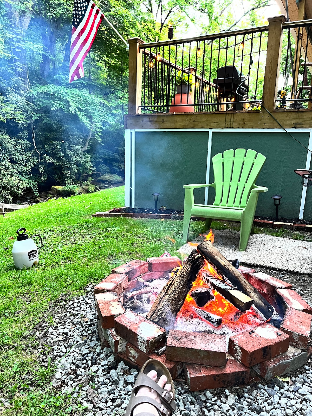A fire pit is surrounded by a circular brick border and filled with glowing logs. Adjacent to the pit, a green Adirondack chair is positioned on a grassy area, with a flag gently waving in the breeze nearby. The outdoor deck can be seen in the background, nestled among lush greenery.