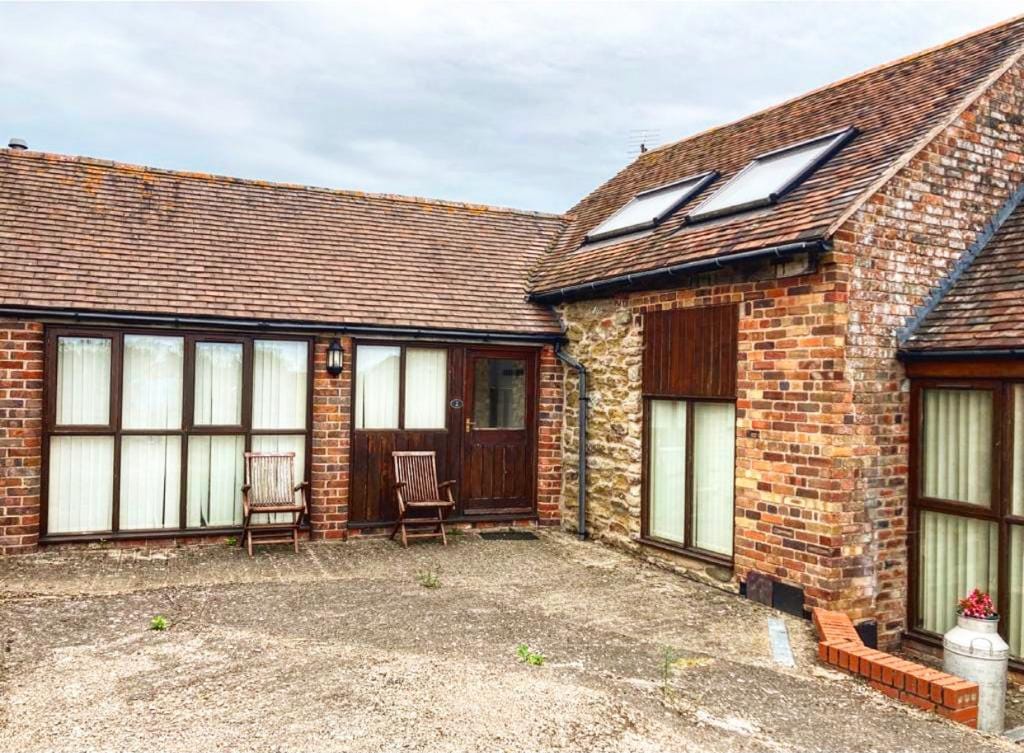 The exterior of the barn conversion is displayed with warm brick walls and a rustic stone accent. Two wooden rocking chairs are positioned on the gravel patio, providing a quiet spot to enjoy the surroundings. Skylights enhance the natural light in the upper level.