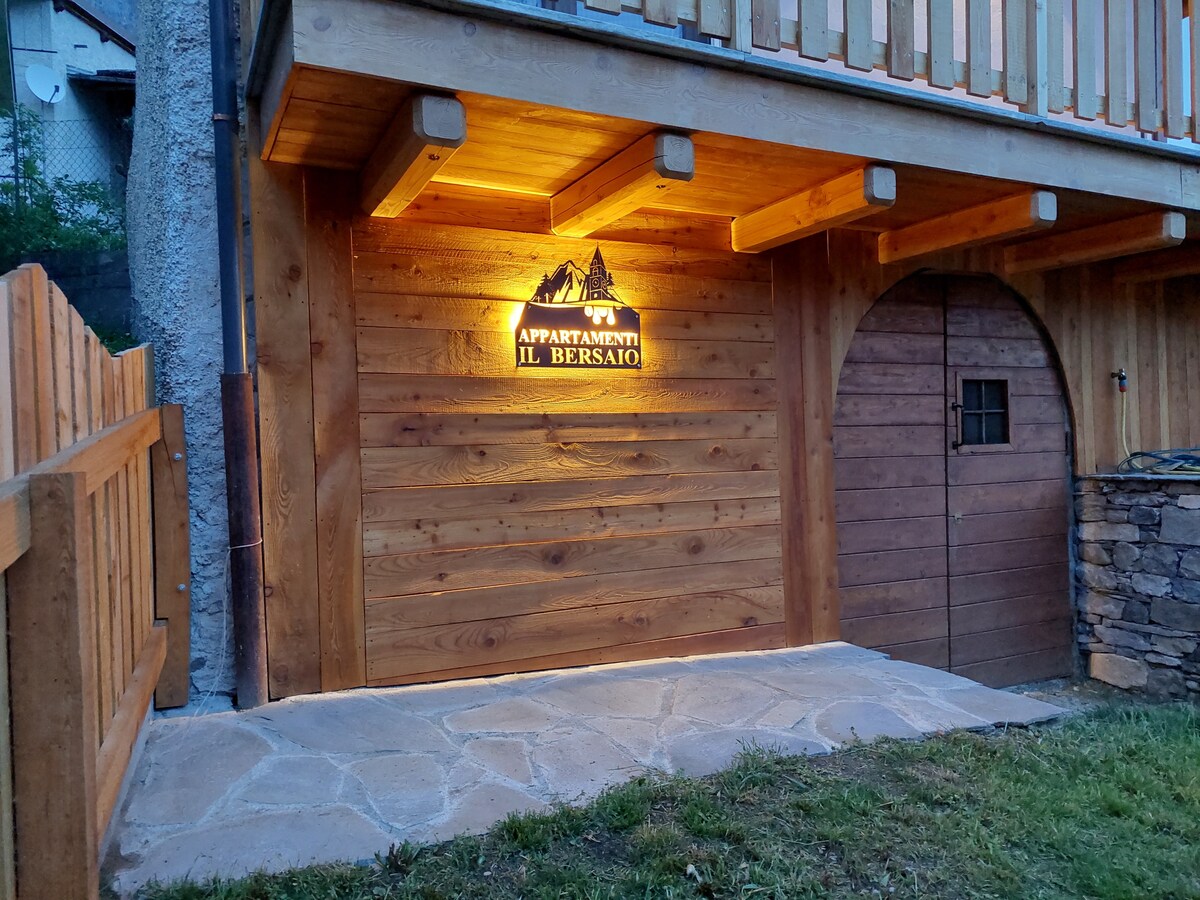 The exterior entrance is highlighted by warm lighting, featuring a wooden facade with the name 'Appartamenti Il Bersaio' prominently displayed. A stone pathway leads to a rustic wooden door, framed by natural surroundings and a wooden fence.