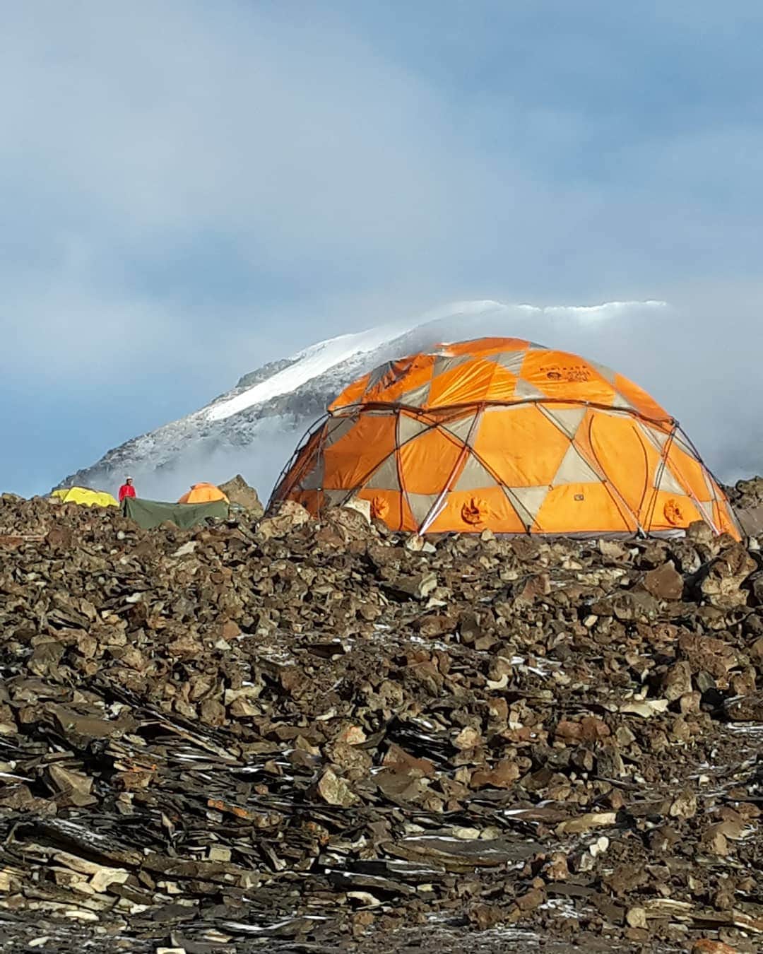 An orange dome-shaped tent is positioned among rocky terrain, with a mountain peak visible in the background, partially covered in snow. The sky appears bright and cloudy, suggesting an adventurous atmosphere, with additional tents in muted colors scattered nearby.
