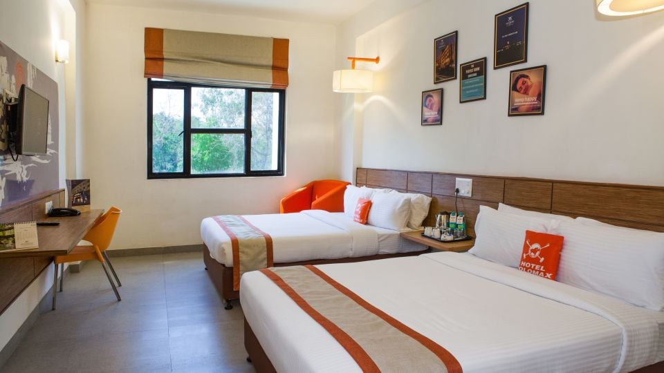 A well-furnished guest room is presented with two spacious beds, minimalistic bedding, and a modern desk. A vibrant orange seating area adds a pop of color. Natural light filters through a large window, enhancing the welcoming ambiance.