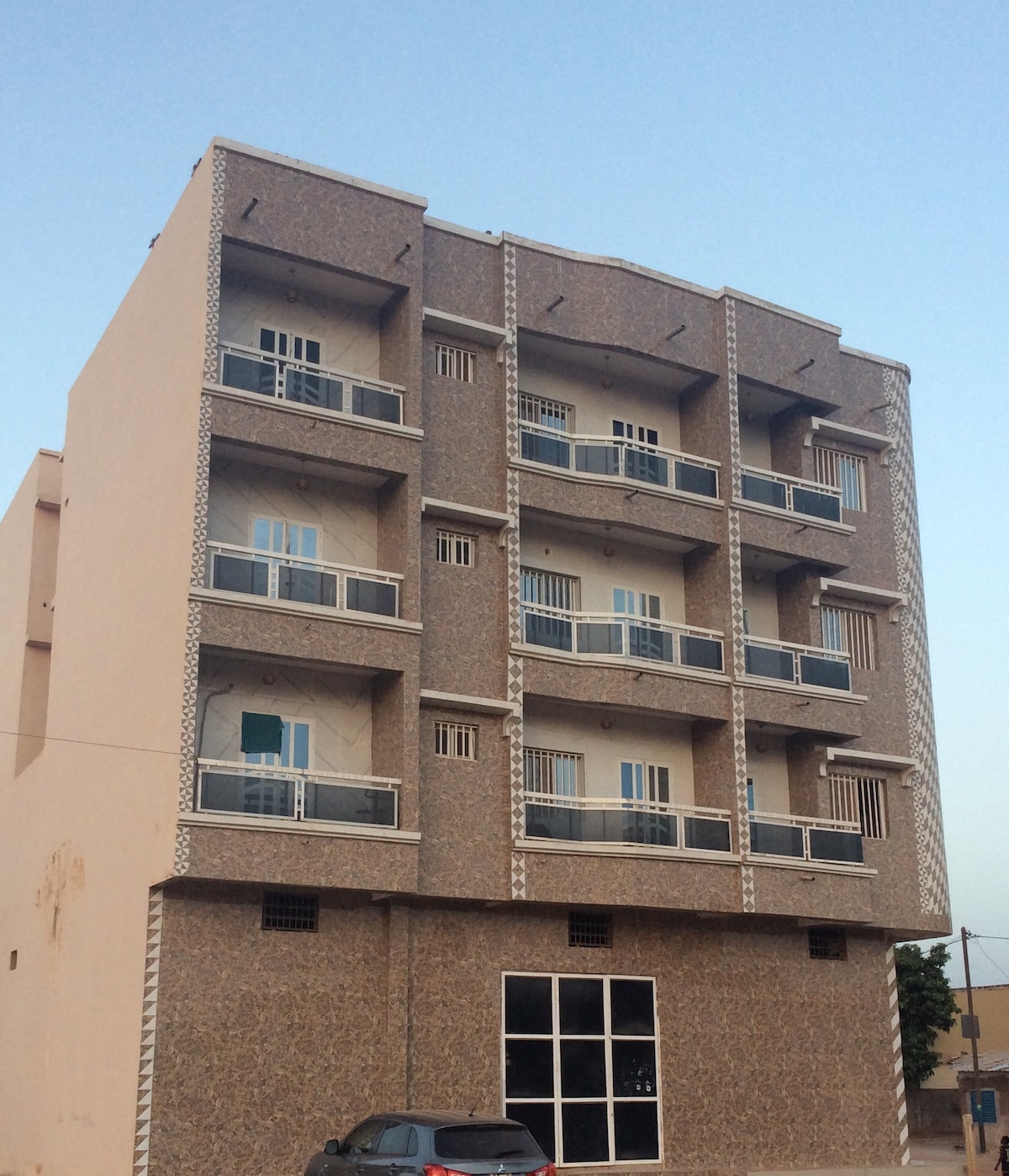 The exterior of a four-story building is showcased, featuring a textured facade with decorative elements. Balconies with railings are visible on each level, while large windows allow natural light into the interior. A parked car is seen in front of the entrance.