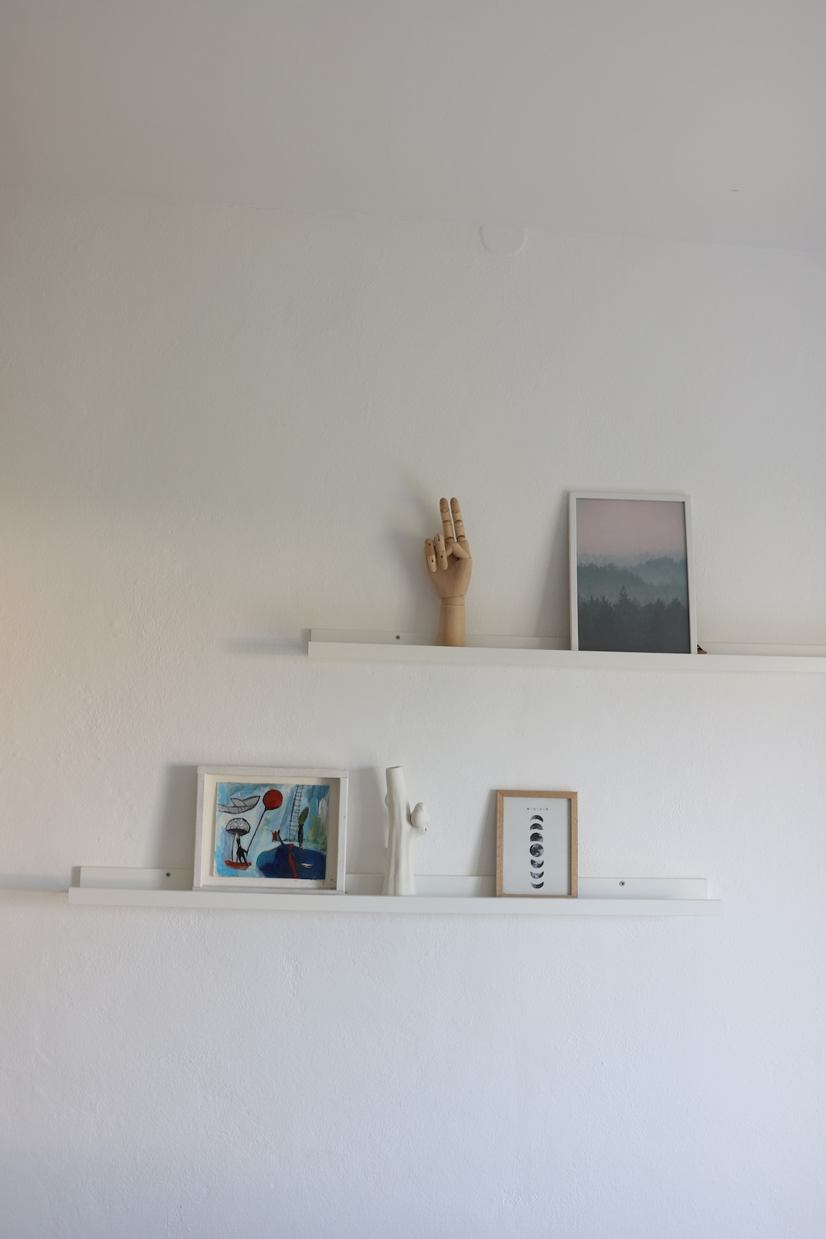 White walls are complemented by two floating shelves, showcasing a variety of decorative items. A wooden hand sculpture gestures upwards, while framed artwork featuring soft colors and abstract designs is displayed, creating a blend of personal touches and artistic expression.