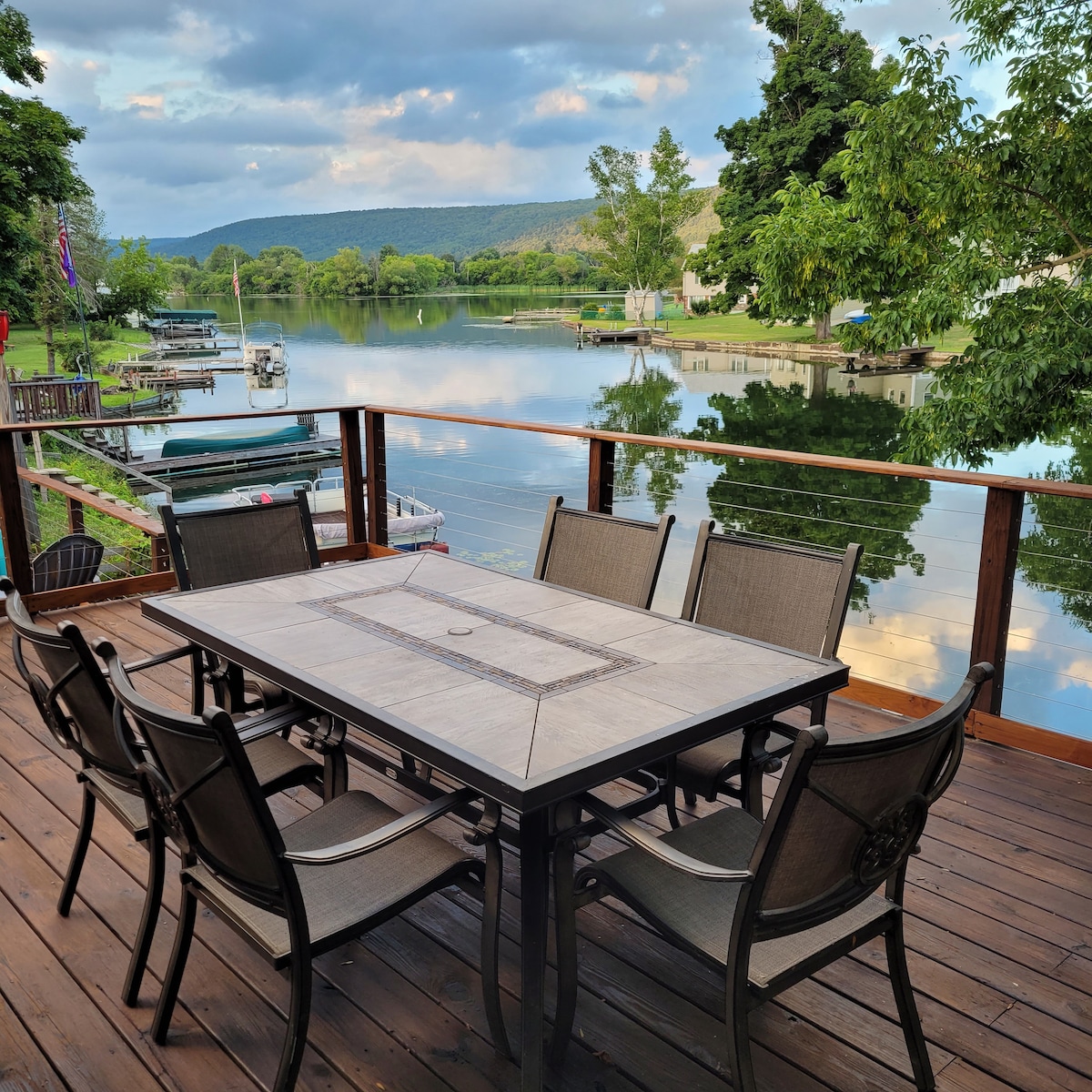 A dining table is set on a wooden deck with seating for six, overlooking a calm lake. The serene water reflects surrounding trees and distant hills. A gentle breeze stirs the atmosphere, framed by clouds in a soft blue sky.