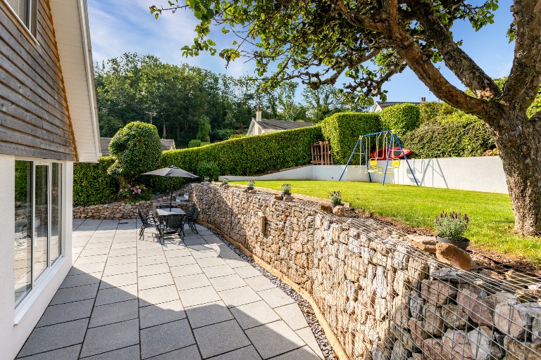 A spacious patio area is framed by a stone wall and features a round dining table with several chairs. A shaded umbrella provides cover, while a swing set is visible in the green lawn beyond, surrounded by neatly trimmed hedges.