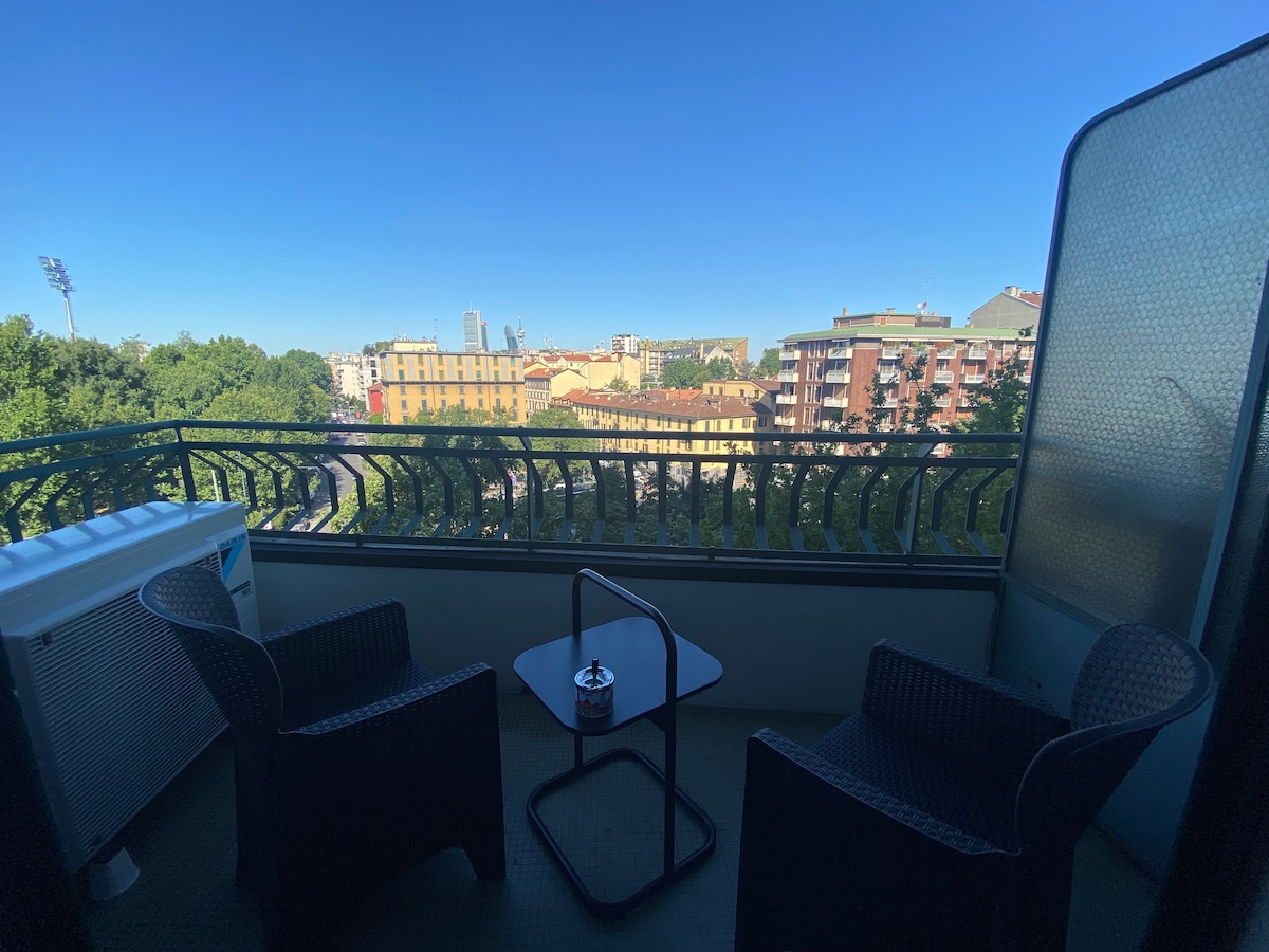 A balcony view is showcased, featuring two wicker chairs positioned around a small table. The background includes a vibrant cityscape, with green trees and buildings visible under a clear blue sky.