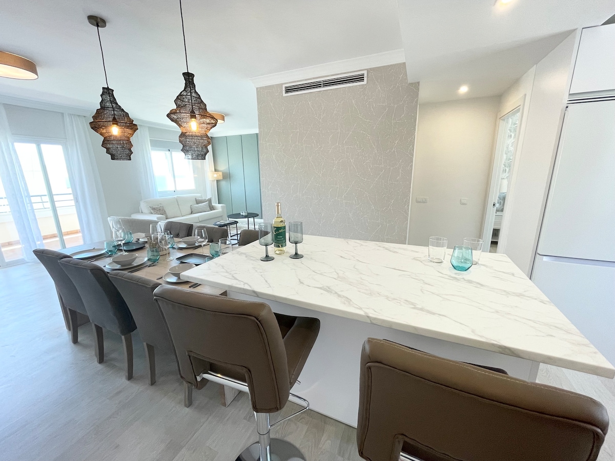 A modern dining area features a marble-top bar with high stools, awaiting guests. Above, unique pendant lights provide soft illumination. A dining table is set for multiple guests, with glassware arranged neatly. The open layout connects to a bright living space with large windows.