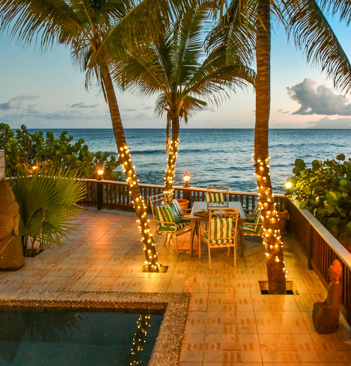 An outdoor dining area is positioned beside the sea, featuring a table surrounded by striped chairs. Palm trees adorned with twinkling lights frame the scene, while a tranquil pool reflects the sunset sky, creating a serene ambiance with gentle waves in the background.
