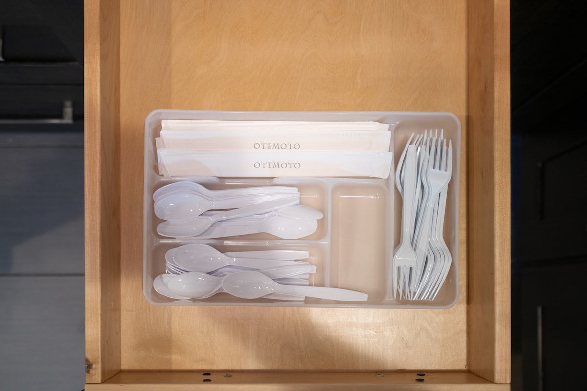 A kitchen drawer is organized with a container holding multiple white forks, spoons, and knives. Plastic cutlery is neatly arranged alongside paper napkins, all easily accessible for dining needs.
