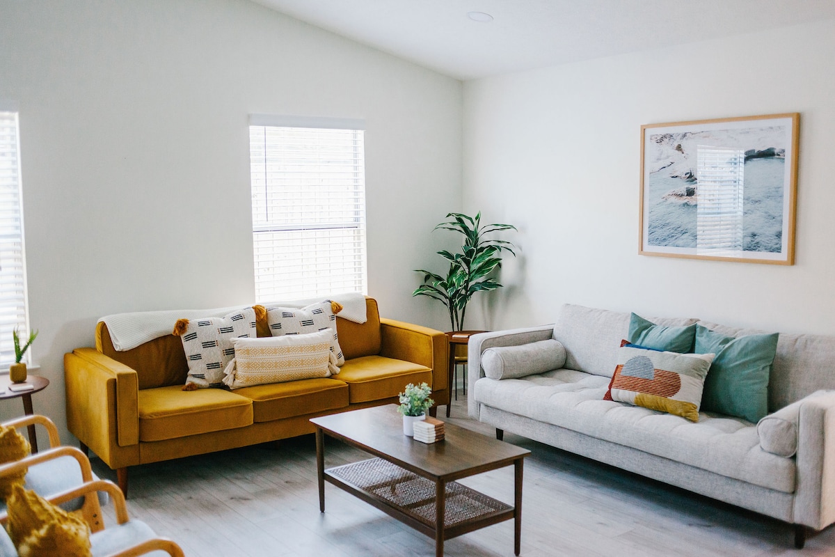 A bright living area is presented, featuring a yellow sofa and a light gray couch positioned around a wooden coffee table. Two windows provide natural light, while a potted plant adds a touch of greenery to the space. Soft decorative pillows enhance the cozy feel.