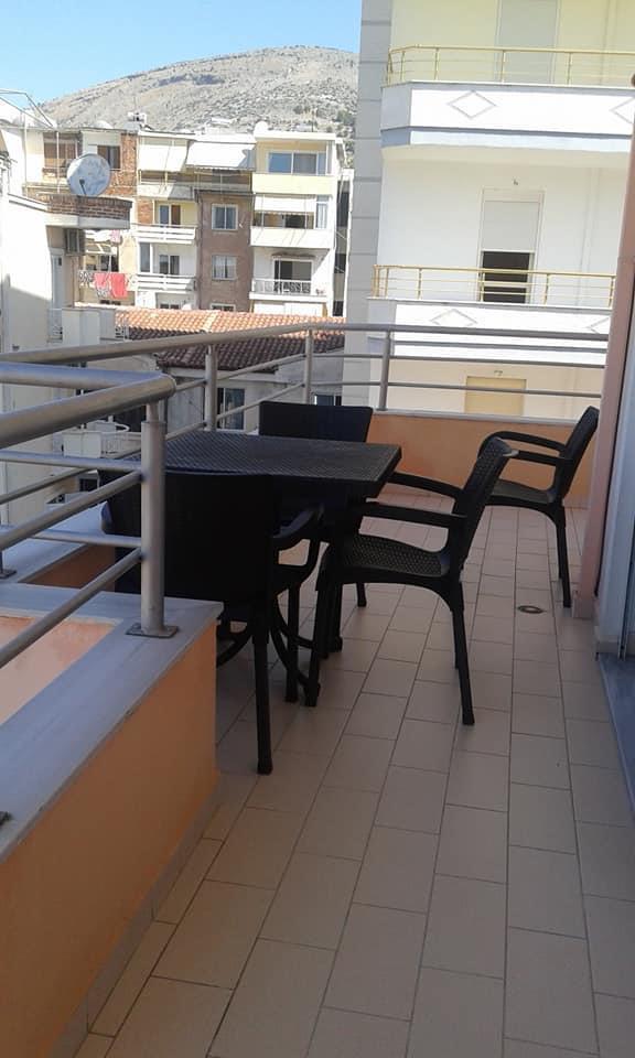A private balcony is shown, featuring a small table accompanied by four chairs. The tiled flooring provides a smooth surface, with a railing offering a view of neighboring buildings and hills in the background.