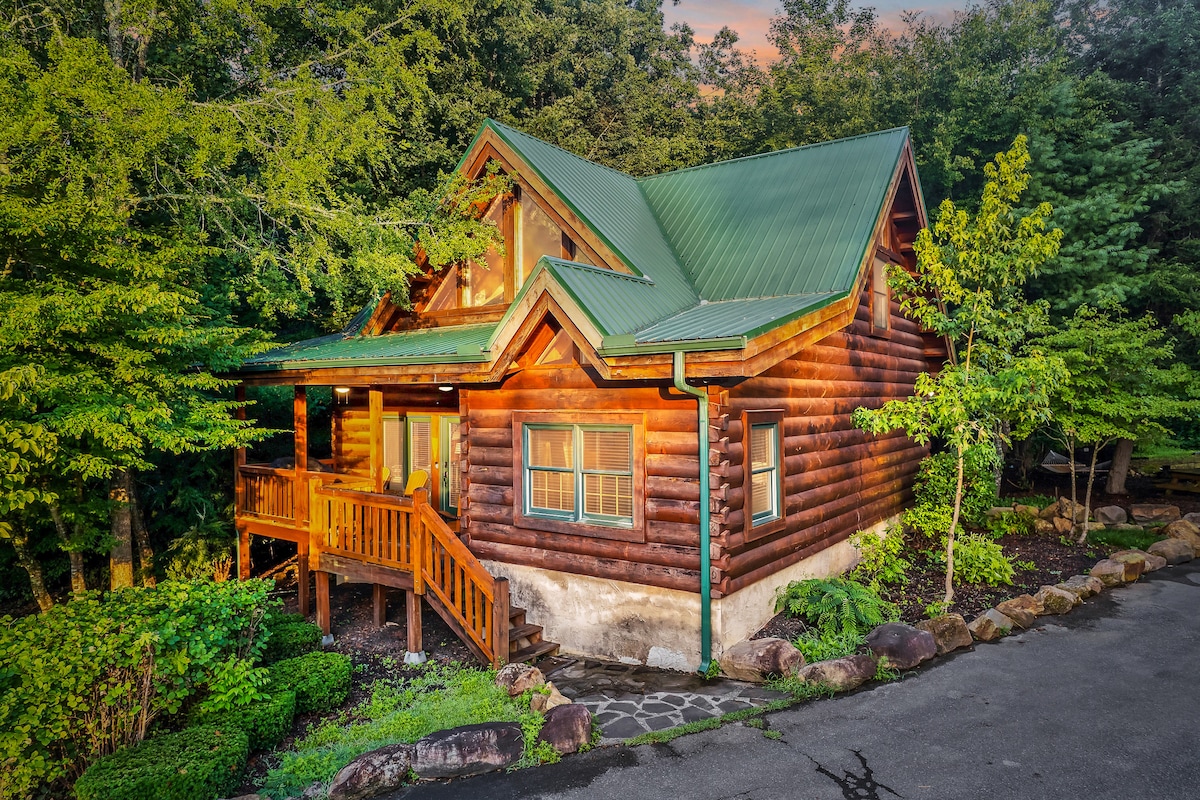A rustic log cabin is depicted surrounded by lush greenery. The sloping green roof complements the rich brown logs, while a spacious porch invites outdoor relaxation. Stone pathways and landscaped gardens enhance the cabin's natural setting, creating a welcoming atmosphere amid the forest.