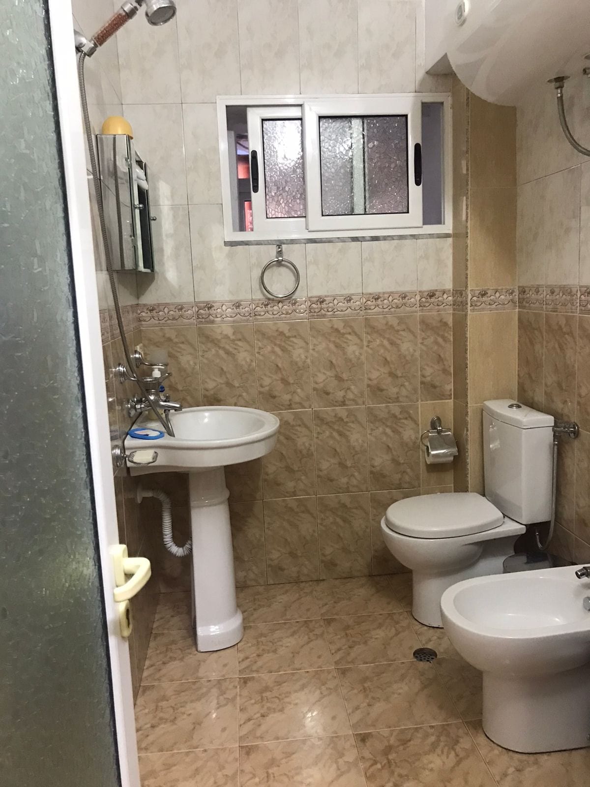 A compact bathroom is presented, featuring a white sink and a toilet alongside a bidet. Smooth, beige tiles cover the wall and floor, giving a clean and modern appearance. A window allows natural light, and a shower is enclosed by a clear glass door.