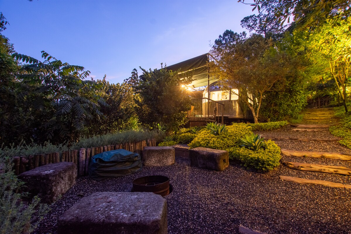 A clear path leads towards the Airstream, surrounded by lush greenery. Evening lights create a warm glow, highlighting the outdoor seating area with stone benches and a fire pit. The serene setting encourages relaxation amidst nature, offering a peaceful retreat.