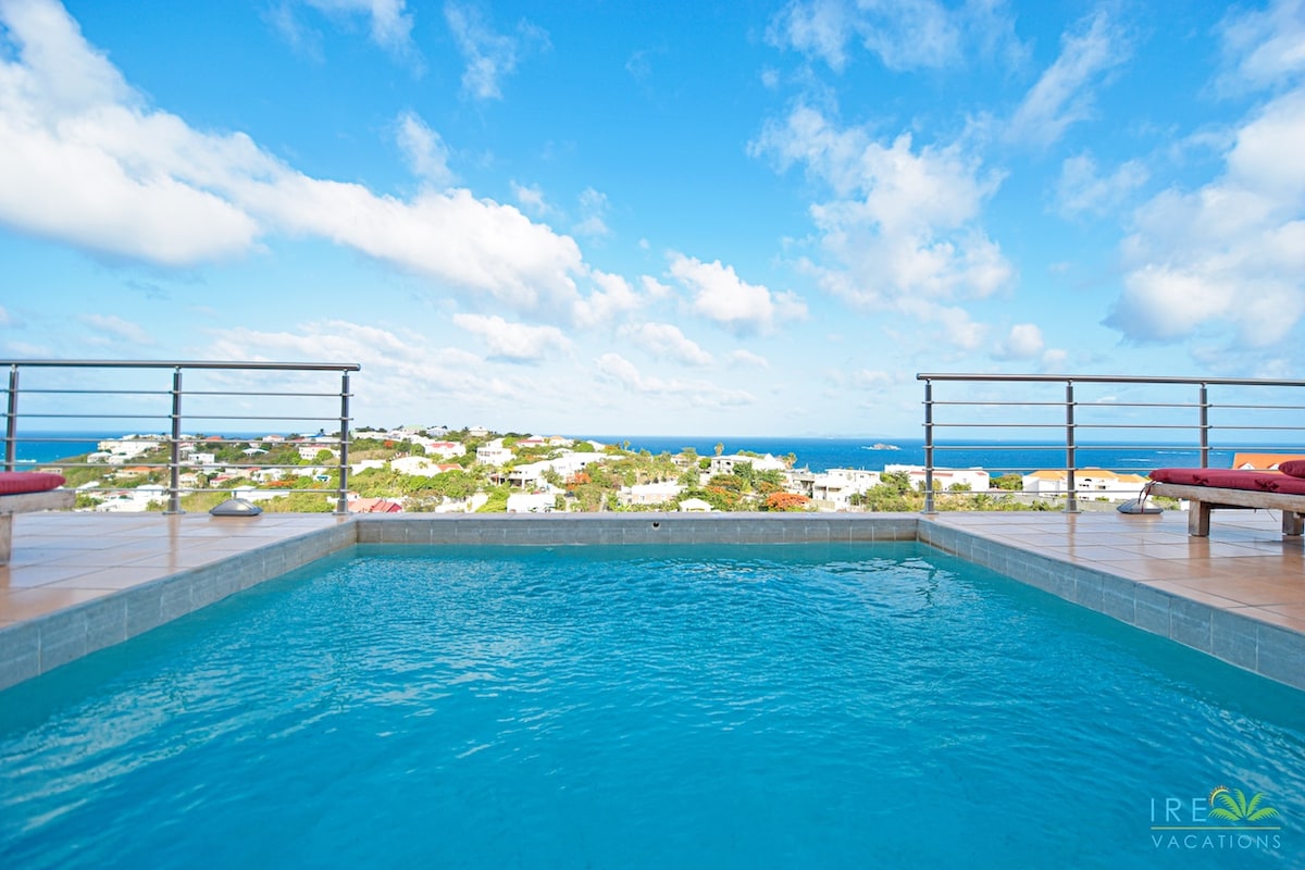 A private pool overlooks lush greenery and the ocean, framed by a clear sky with scattered clouds. Comfortable lounge chairs are positioned at the pool's edge, inviting relaxation while enjoying the expansive views.