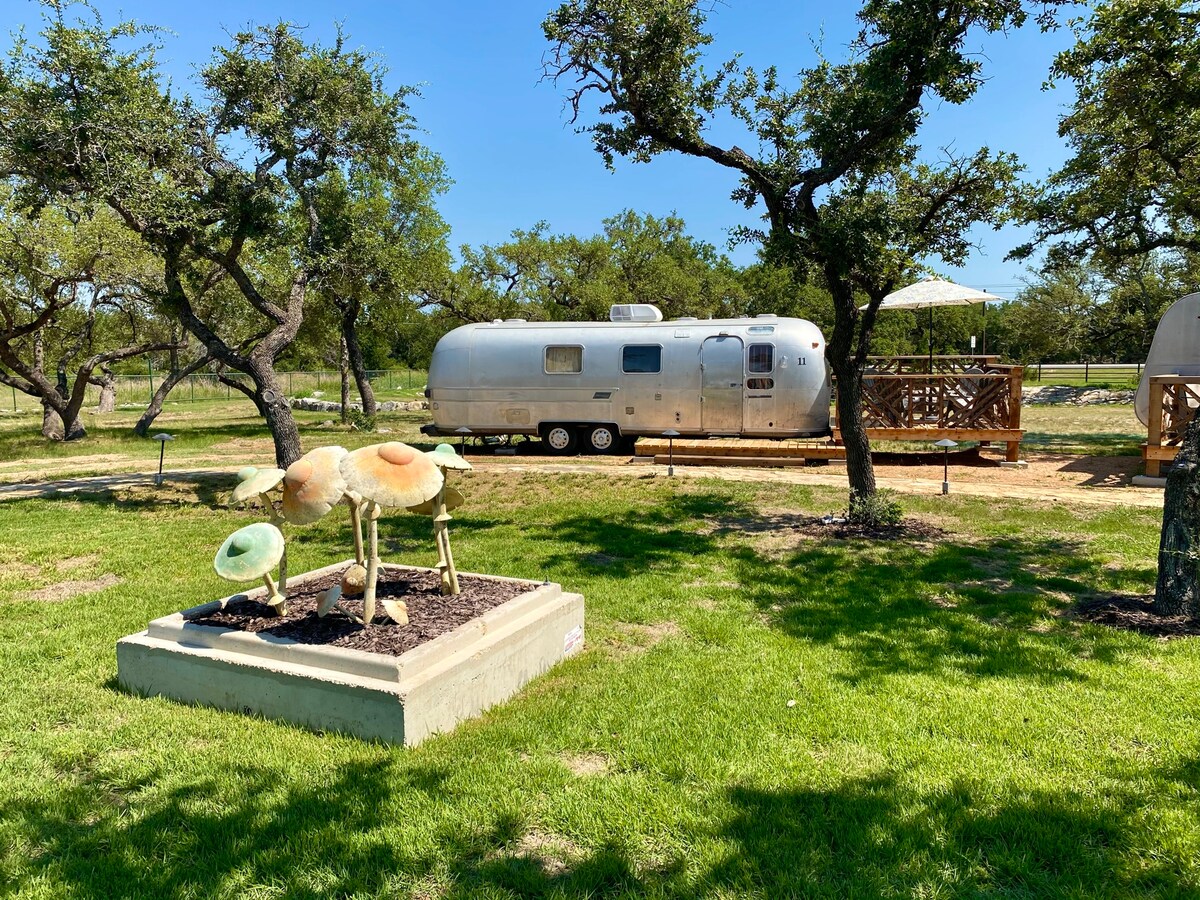 An Airstream trailer is positioned on a grassy area, surrounded by trees. A whimsical sculpture of oversized mushrooms is showcased in the foreground. A wooden deck with an umbrella is visible adjacent to the trailer, inviting relaxation under the shade.