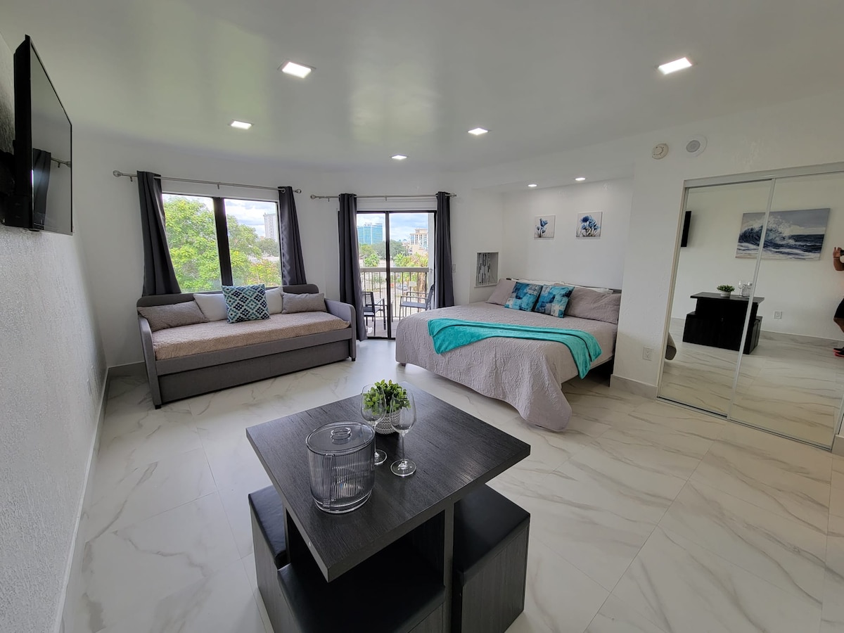 A bright studio features a spacious bed with soft bedding, complemented by a cozy sofa. Large windows allow natural light to fill the room, while a small table with decorative accents sits in the center. Mirrored closets enhance the room's openness.