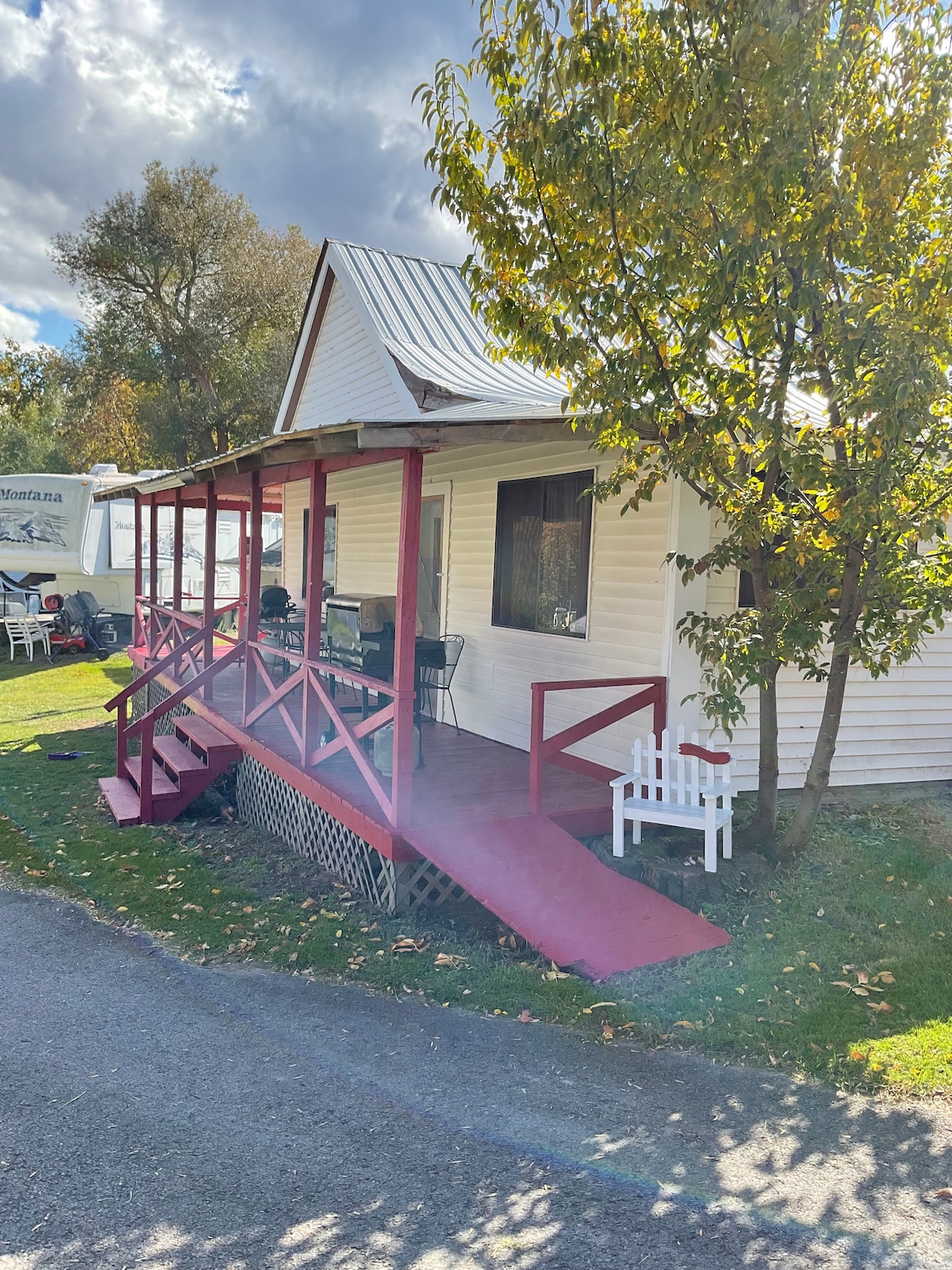 A charming cabin features a welcoming red porch and stairway, providing easy access to the entrance. Surrounding greenery adds a natural touch, with a small white chair positioned on the porch. The cabin's exterior, painted white with a metal roof, adds to its rustic appeal.