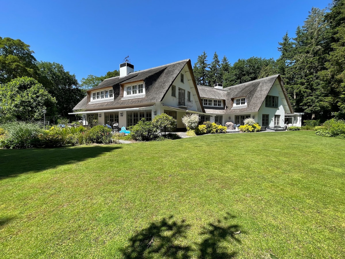 The spacious villa features a thatched roof and is surrounded by lush greenery and well-maintained gardens. Expansive windows reflect natural light, while a manicured lawn invites outdoor activities. The serene backdrop of trees adds to the tranquil setting.