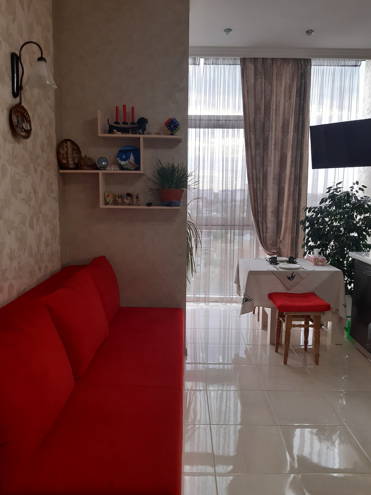 A bright living space features a red couch along a wallpapered wall with decorative shelves. Natural light floods the area, highlighting a small dining table with a chair. Floor-to-ceiling curtains frame the large window, while a potted plant adds a touch of greenery.