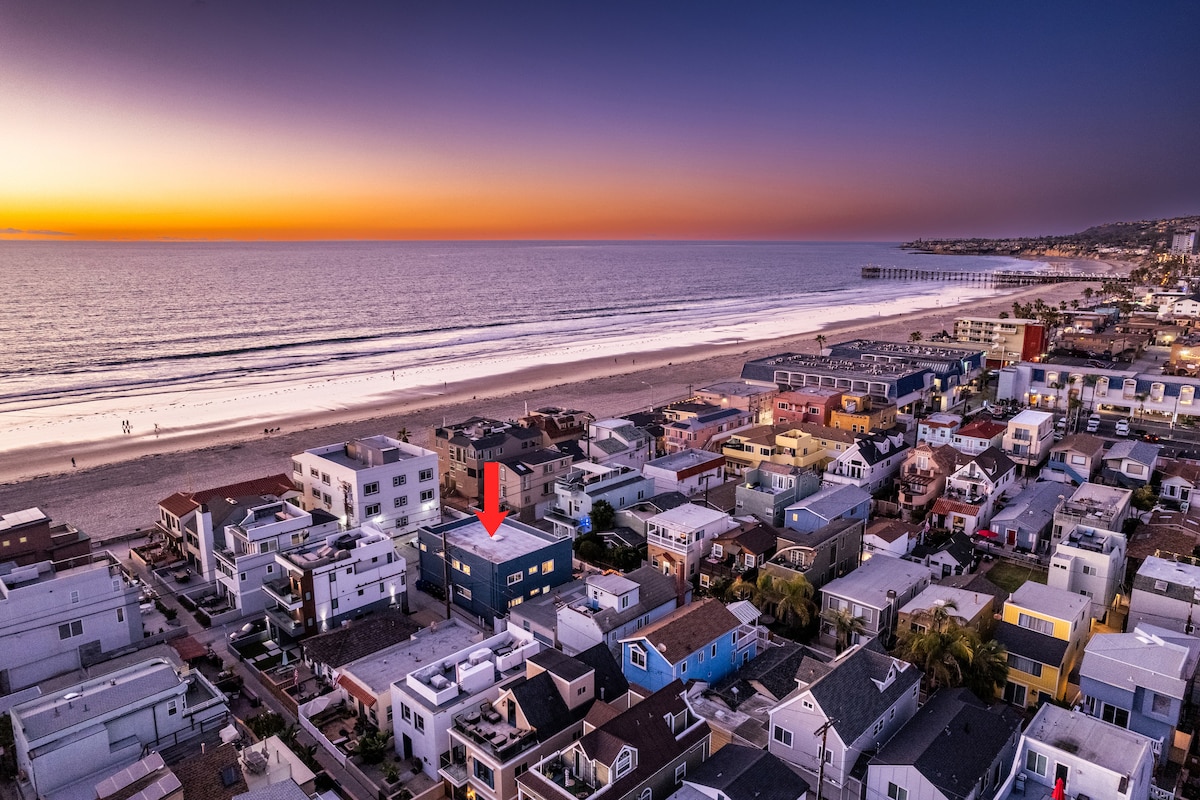 An aerial view captures the beach house located near Mission Beach, with ocean waves gently lapping at the shore during sunset. Colorful rooftops and a vibrant coastline are visible, highlighting the close proximity to the beach and nearby structures.