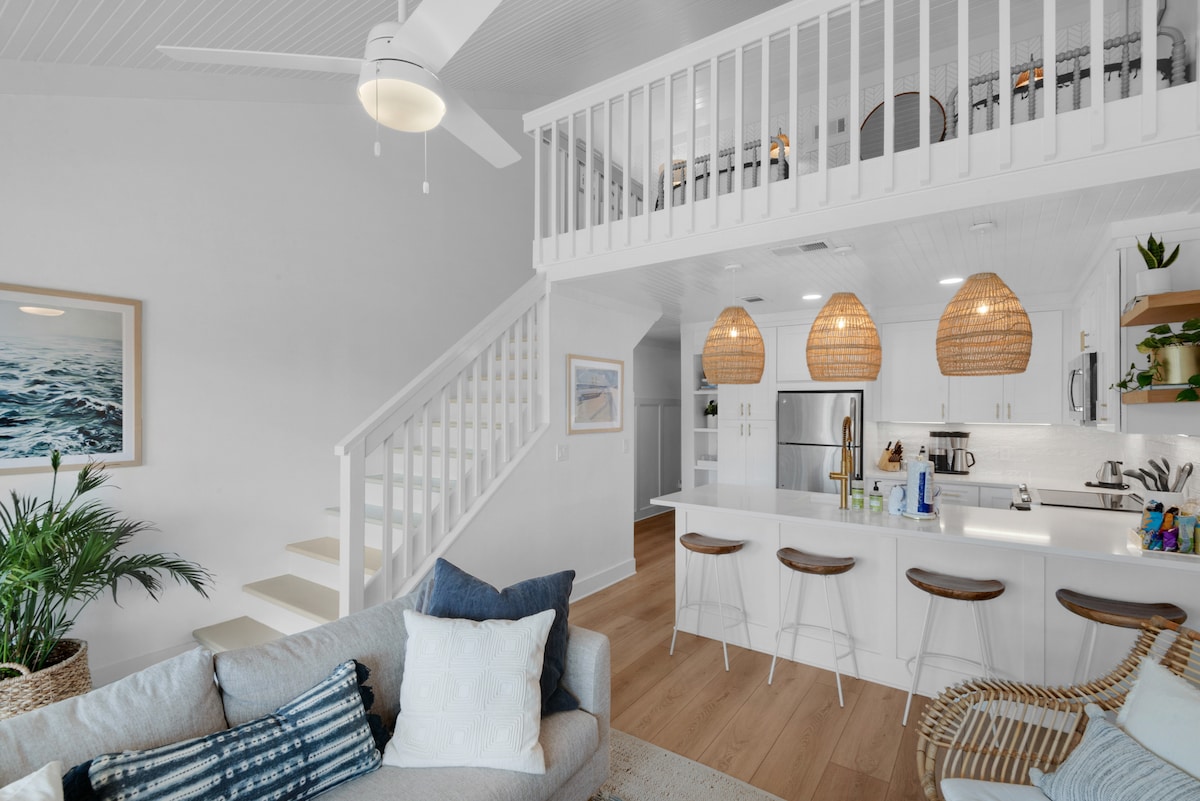 A bright and airy living space features a comfortable sofa with decorative pillows. A staircase leads to a loft area, while woven pendant lights hang above a modern kitchen with sleek appliances and a breakfast counter. Tropical greenery adds a touch of nature.